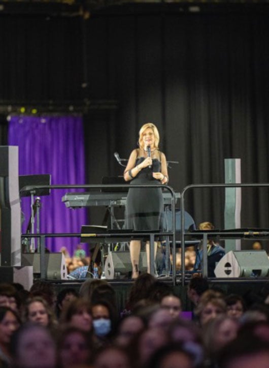 This image presents a scene from a large conference, focusing on a female speaker standing alone on an elevated platform. 